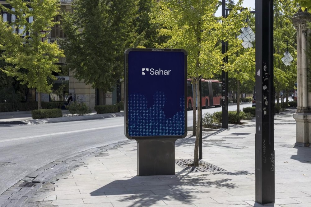 Mobilier urbain JCDecaux sur une avenue plantée : panneau Sahar bleu nuit, logo en tête et trame de points cobalt en pied.